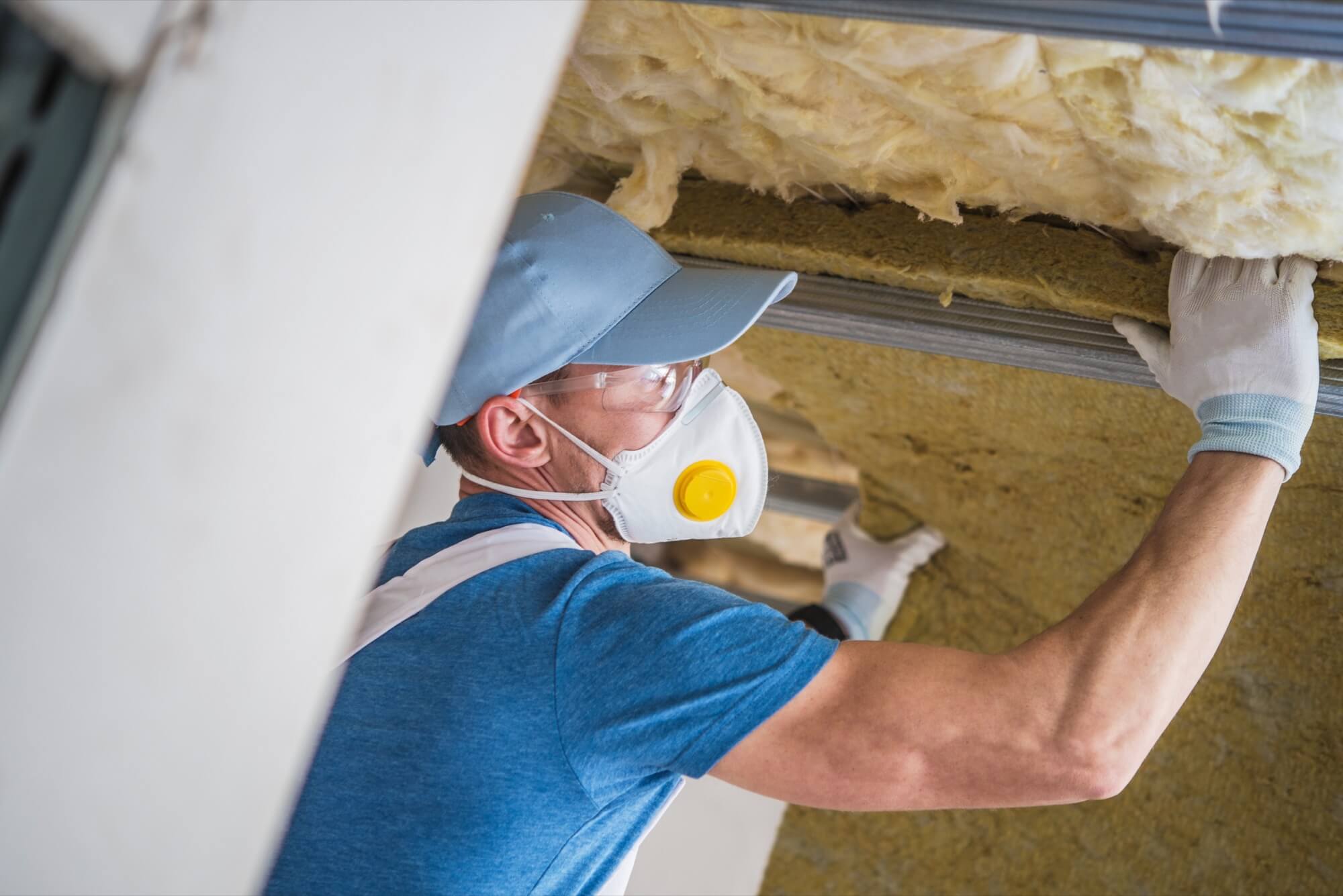 A craftsman wearing a face mask and gloves installs mineral wool in a roof for insulation.